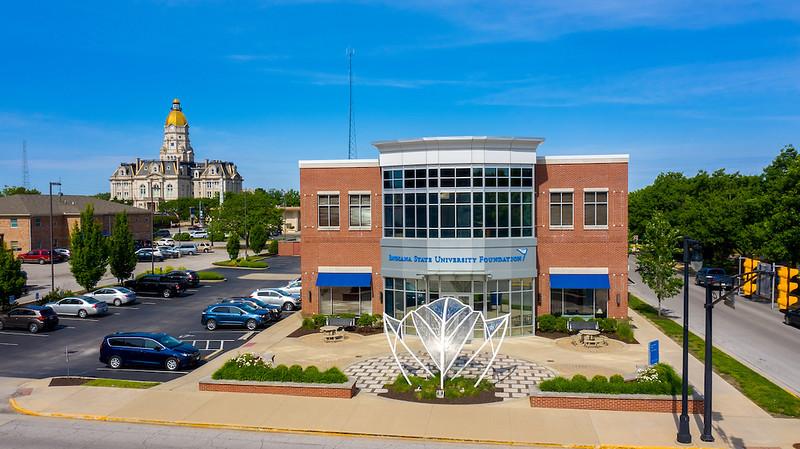 Modern two-story brick building with large windows and blue awnings labeled Indiana State University Foundation, parking lot, and historic courthouse with gold 