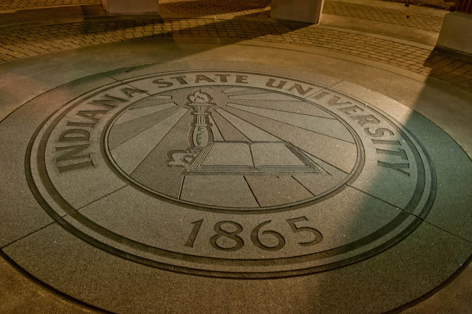 Indiana State University emblem etched in stone with a torch and an open book, dated 1865, illuminated by street lighting at night.