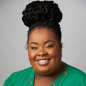 Smiling woman with dark braided hair in a high bun, wearing a green top against a plain gray background.