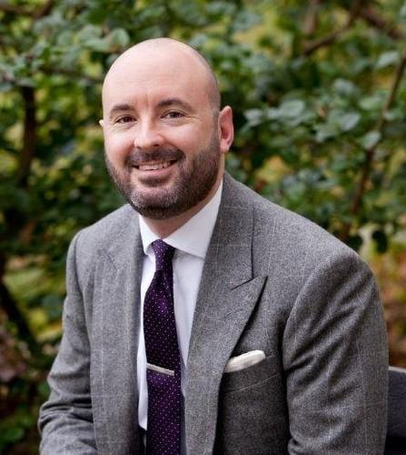 Smiling bald man with beard wearing gray suit jacket, white shirt, and dark tie, seated outdoors with green foliage background.