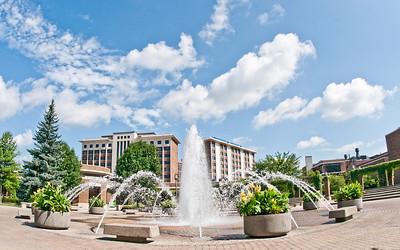 Fountain with multiple water jets in a plaza surrounded by trees and buildings under a blue sky with scattered clouds.