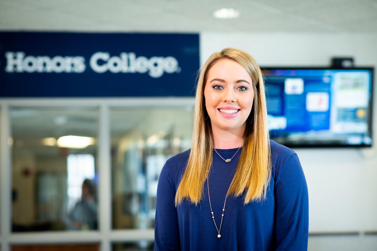 Young woman with long blonde hair wearing a blue top and two necklaces standing indoors in front of a sign that reads Honors College.