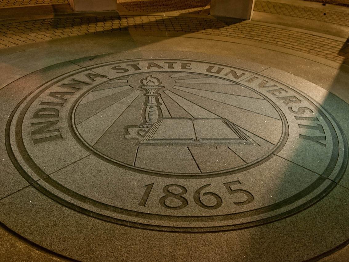 Indiana State University seal embedded in concrete with torch, book symbols, and the year 1865, lit by yellowish outdoor lighting at night.
