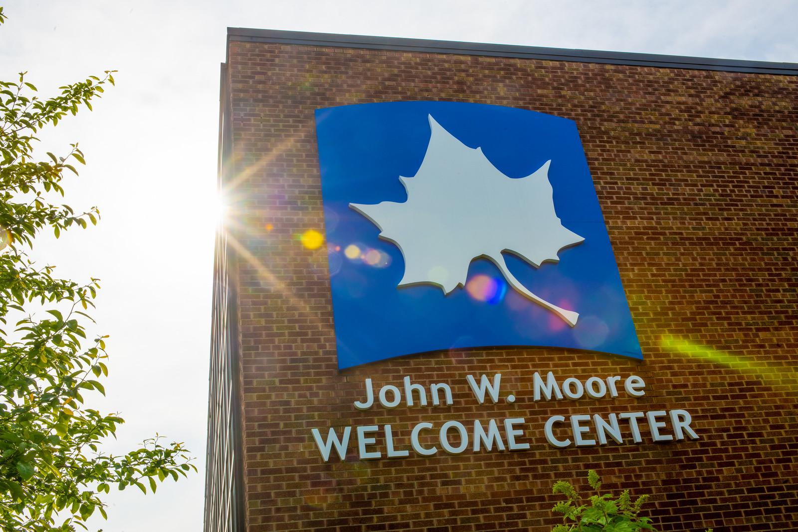 Brick building with a blue sign featuring a white leaf and text reading John W. Moore Welcome Center with sunlight and green leaves nearby.