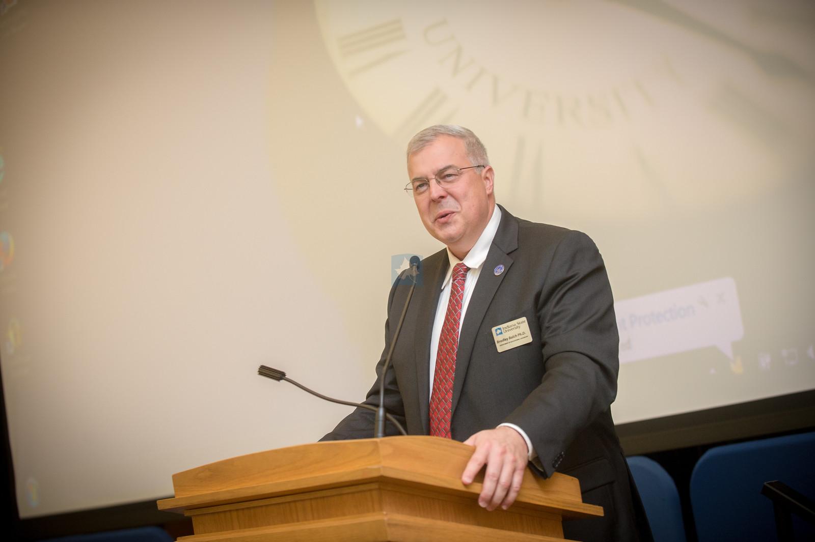 Man in suit and red tie speaking at wooden podium with microphone, university logo projected on screen behind him.