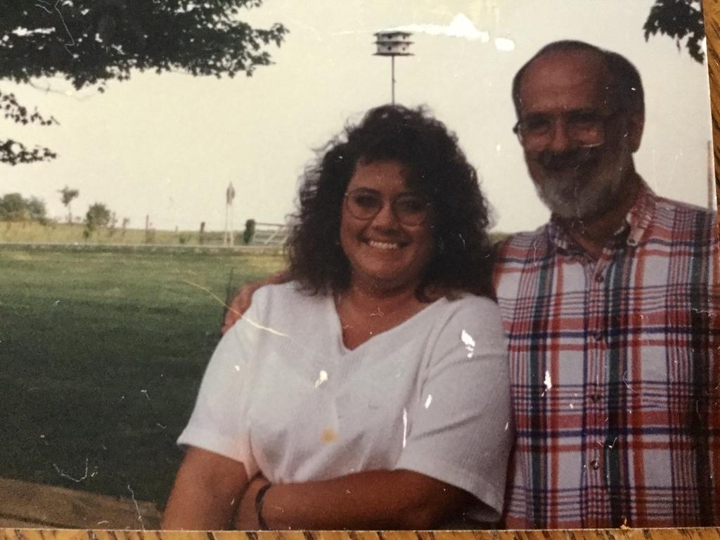 Smiling woman with glasses and curly hair stands outdoors beside older man with glasses and plaid shirt, arm around her shoulder under tree shade.
