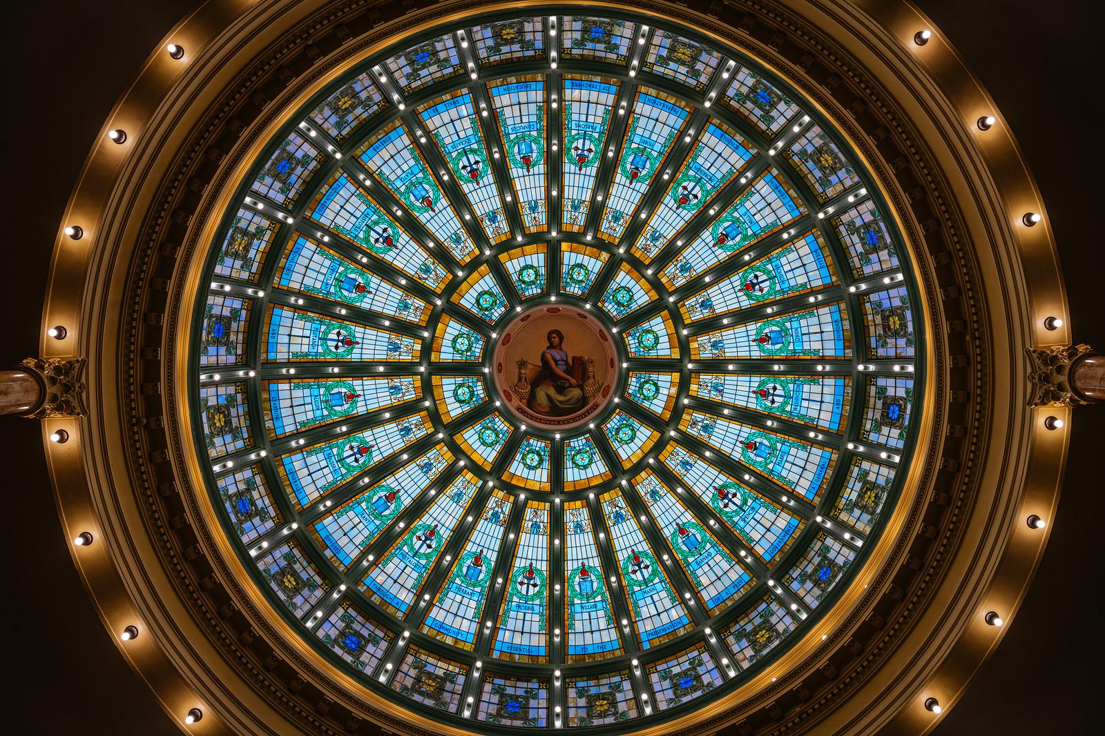 Ornate circular stained glass dome ceiling with bright blue, gold, and green panels and a painted central figure of a seated woman.