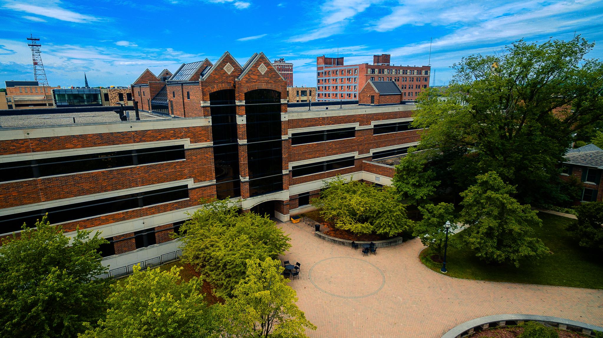 Brick university buildings with large black windows surround a courtyard with trees, outdoor tables, and chairs under a partly cloudy blue sky.
