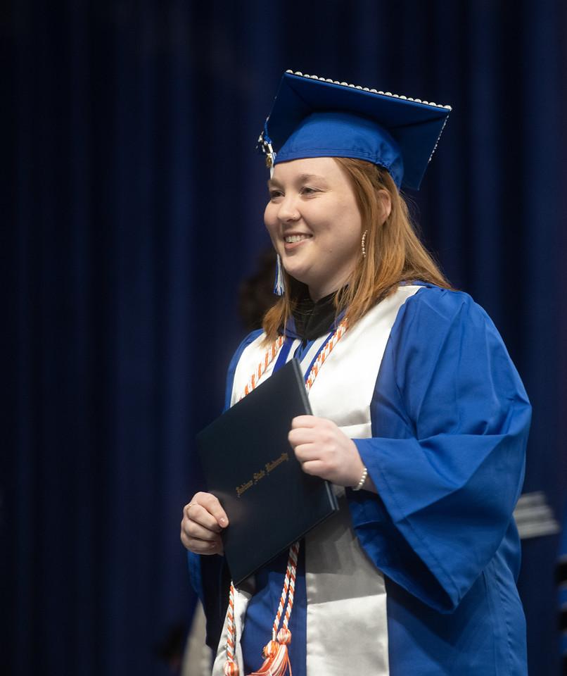 Young woman in blue graduation gown and cap holding diploma folder, smiling against a dark blue curtain background at a commencement ceremony.