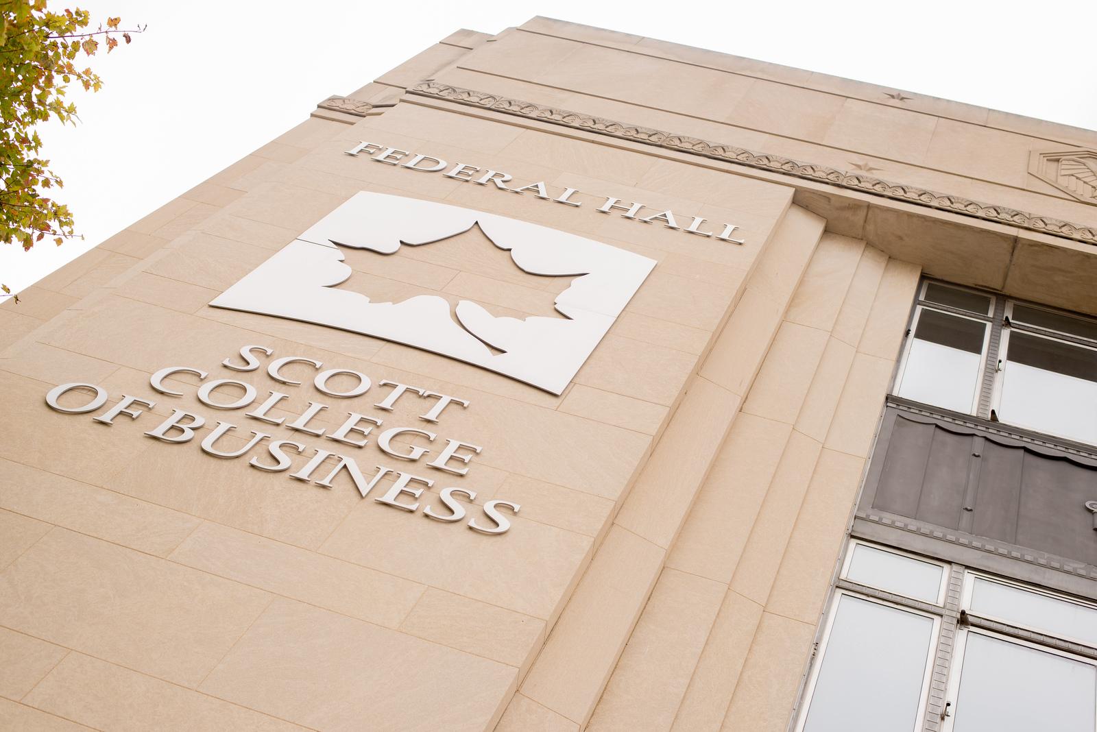 Exterior wall of Federal Hall at Indiana State University with Scott College of Business signage and a leaf logo above windows and a tree branch.