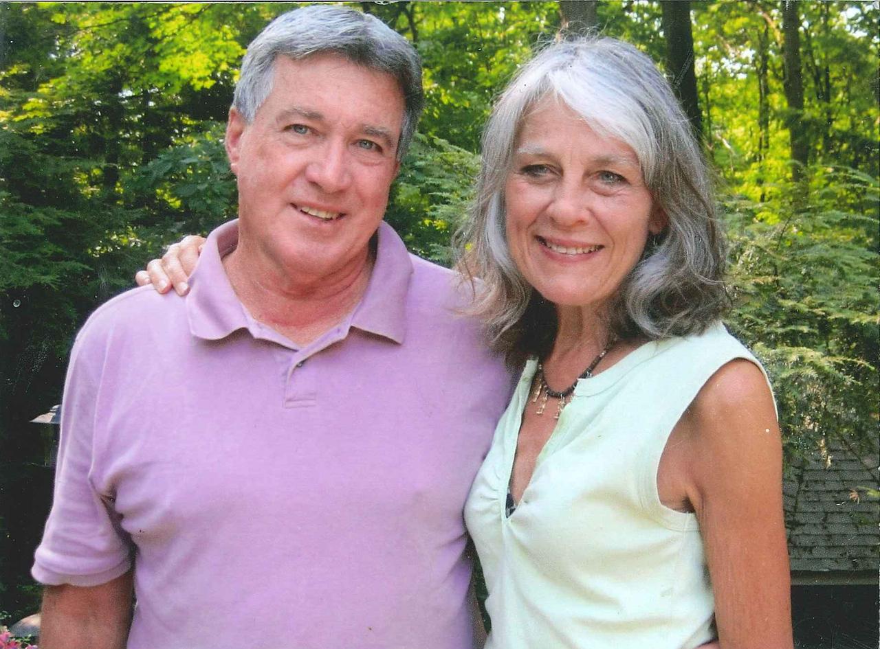 Man in a light purple polo shirt and woman in a sleeveless light green top smiling outdoors with green trees in the background.