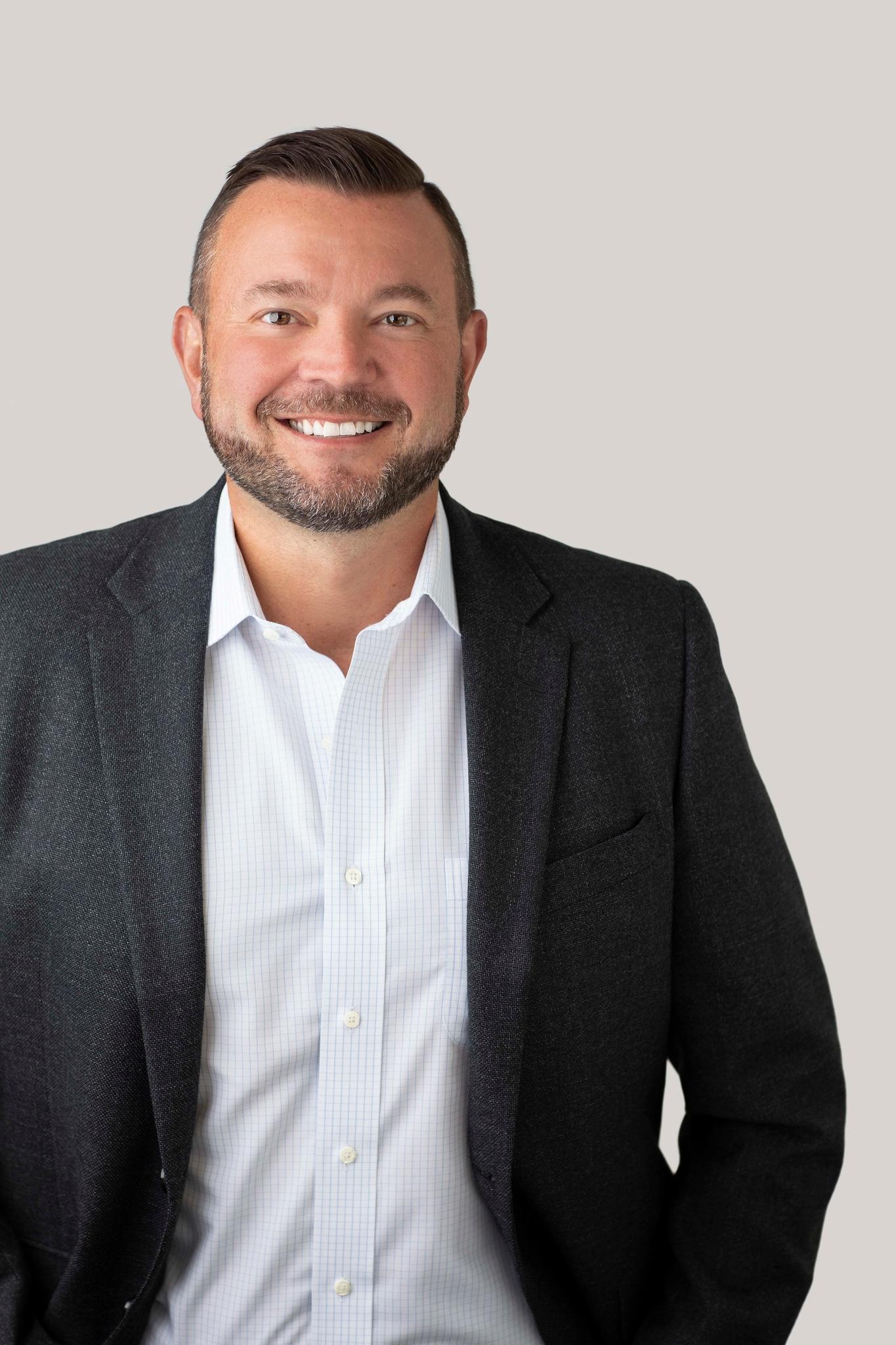 Smiling man with short hair and beard wearing a dark blazer and white checkered shirt against a plain neutral background.