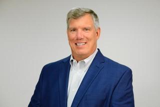 Man in blue blazer and white shirt smiling against a plain light gray background.