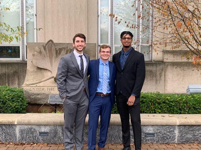Three young men in suits smiling and standing side by side outdoors in front of a stone wall with greenery.