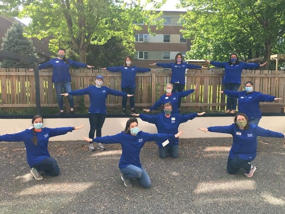 Group of eleven masked adults wearing matching blue shirts standing and kneeling outdoors with arms outstretched to maintain social distance.