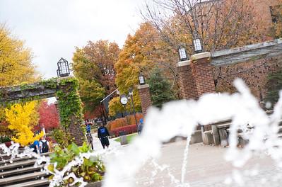 Fountain water spraying in front of a brick archway and walkway lined with autumn trees and a few people walking on campus.