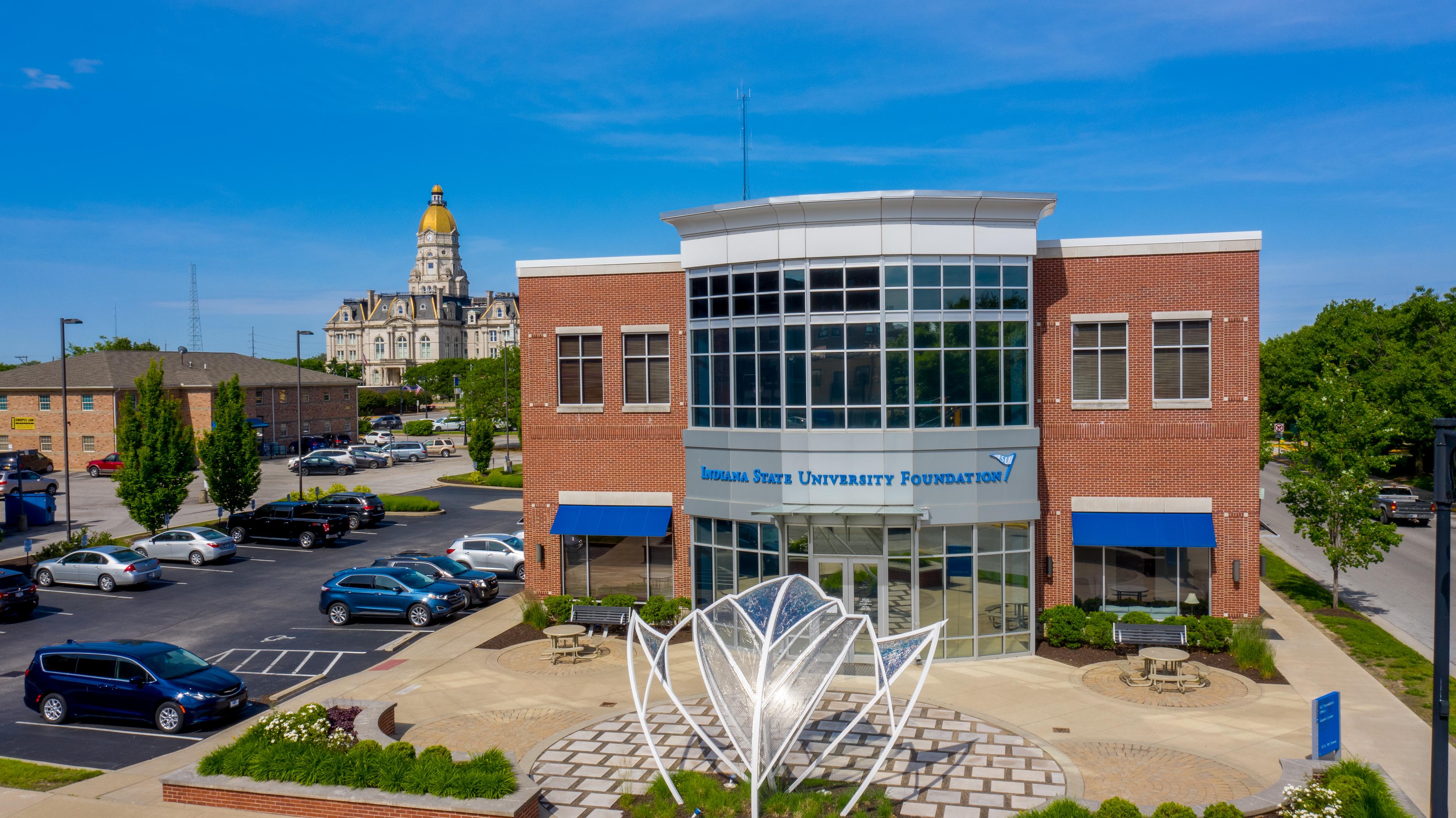 Indiana State University Foundation building with blue awnings, a metal flower sculpture in front, parked cars, and a historic courthouse with a gold dome in th