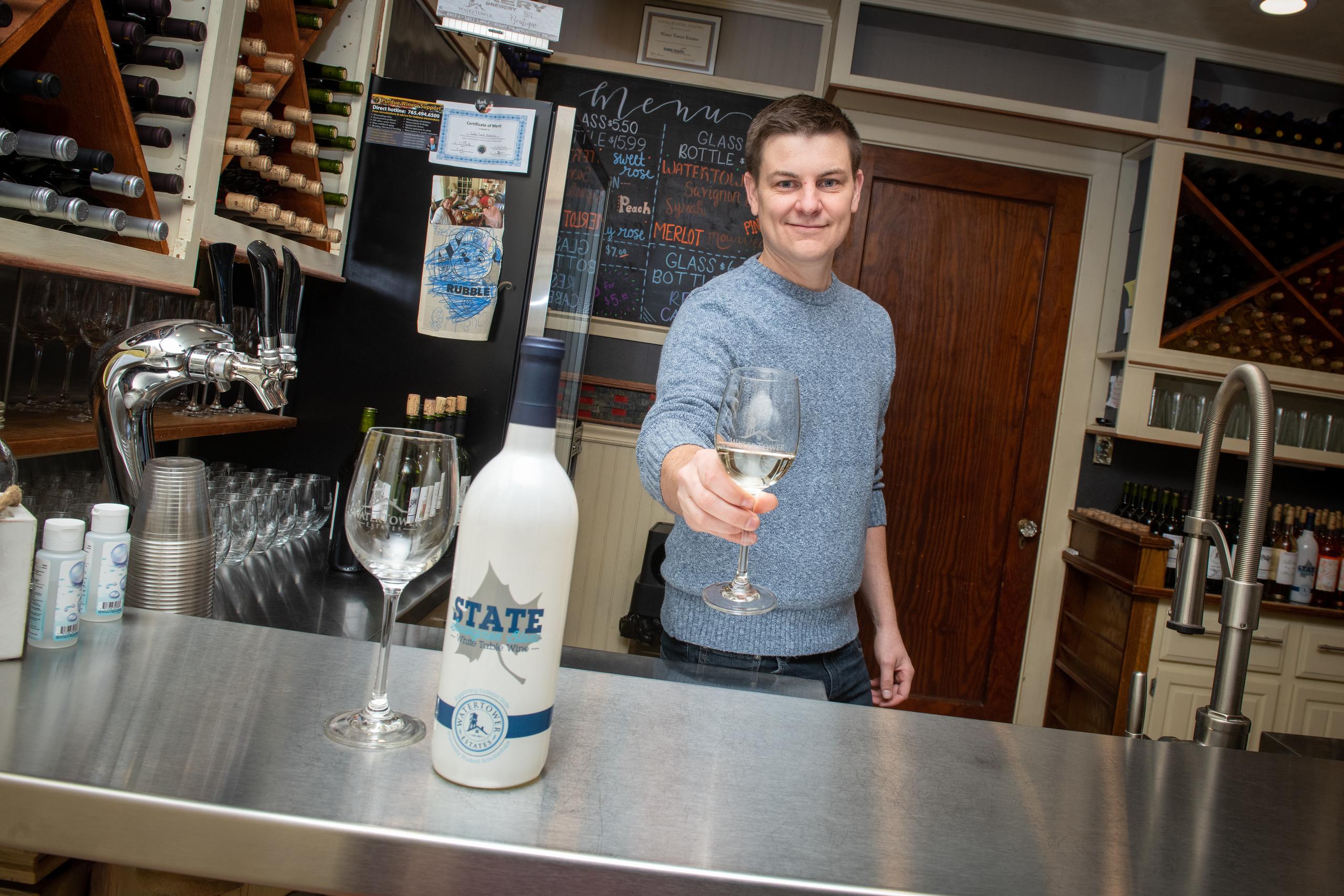 Man in gray sweater behind bar holding out a glass of white wine, with a bottle labeled State White Table Wine and an empty wine glass on the counter.