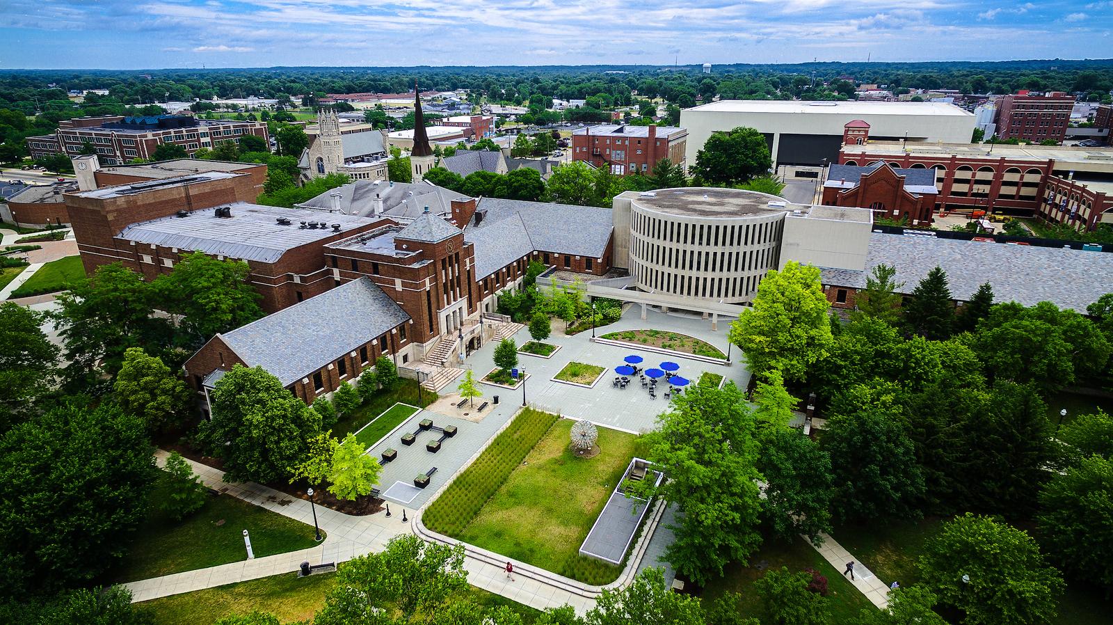 Aerial view of Indiana State University campus with brick buildings, walkways, green trees, and outdoor seating with blue umbrellas under a partly cloudy sky.