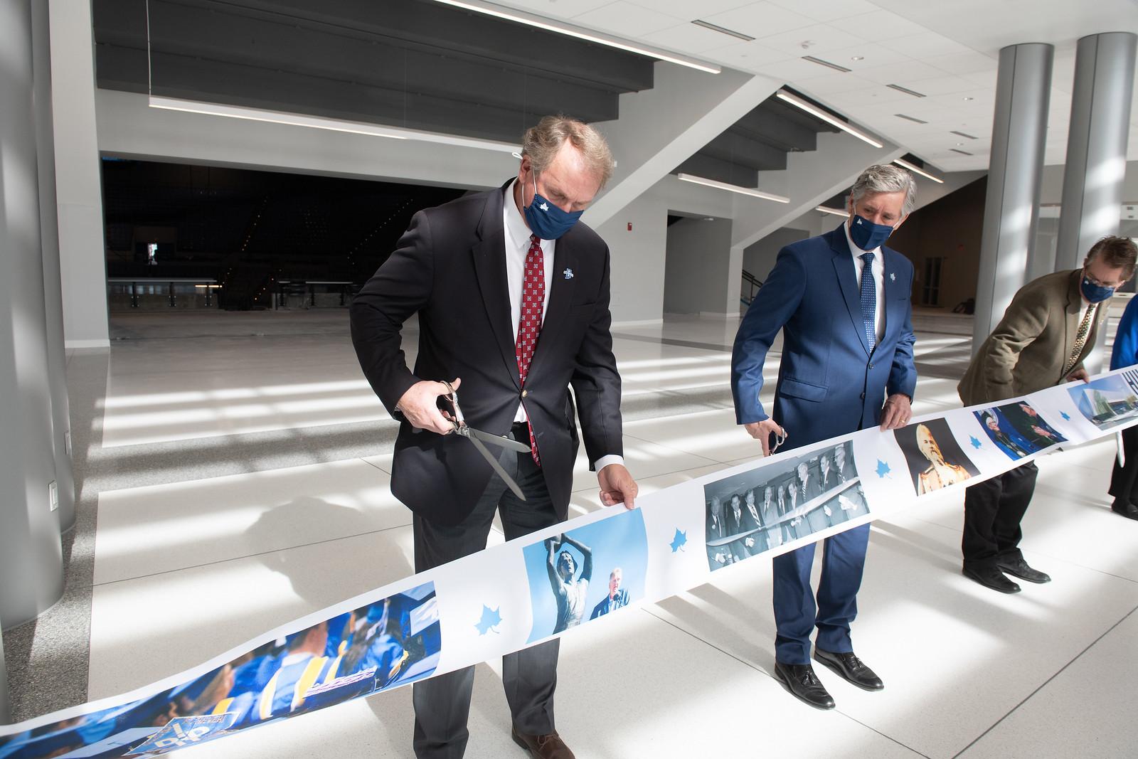 Four men in suits and masks cut a long printed ribbon inside a bright modern building during a ribbon-cutting ceremony.