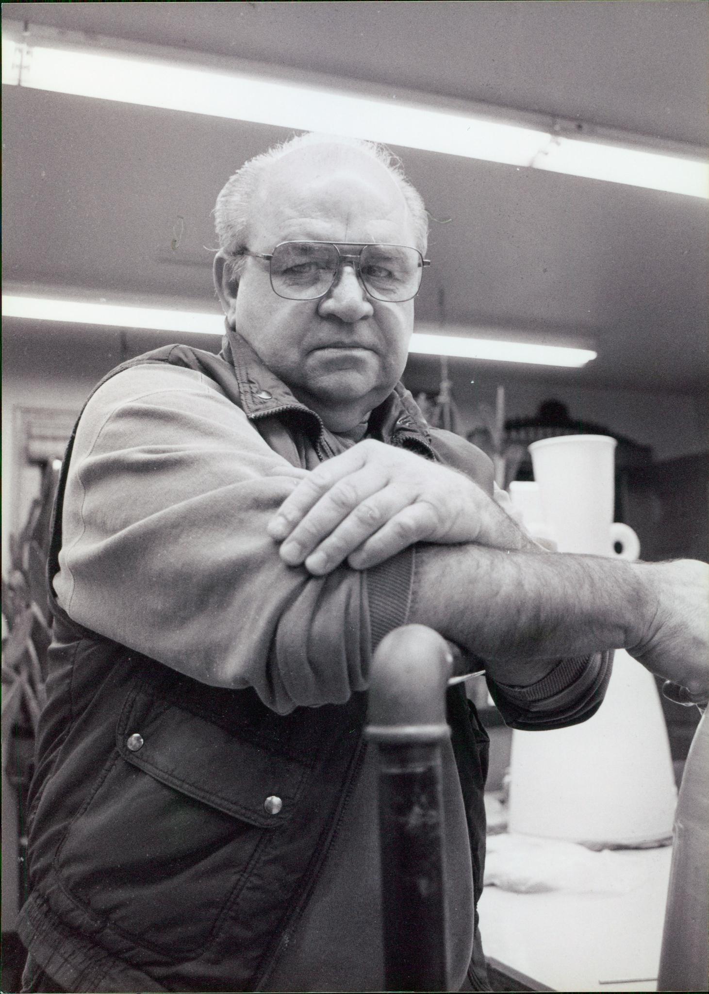 Older man with glasses and receding hairline wearing a jacket, leaning on a table with pottery and fluorescent lights overhead in a workshop.