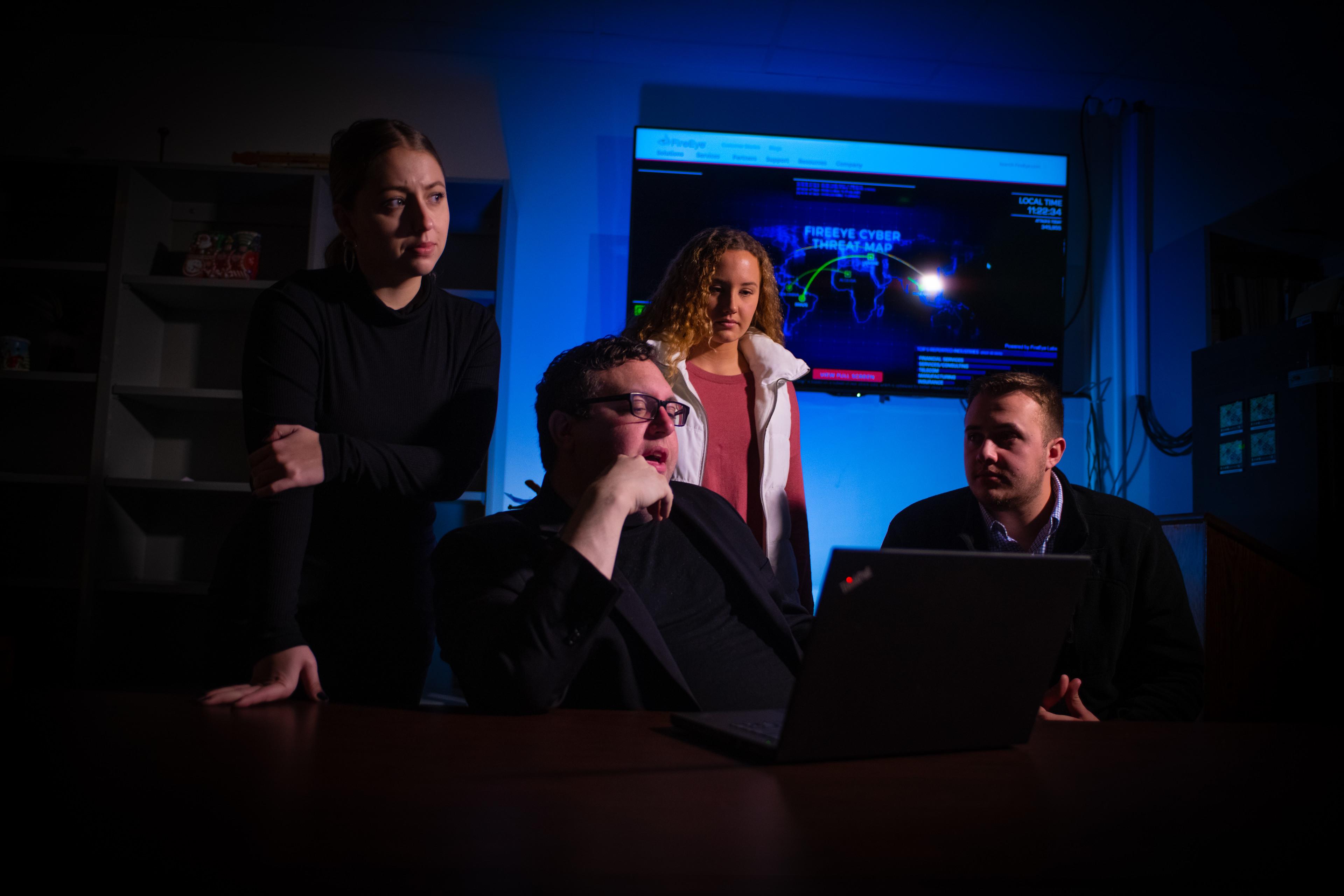 Four people gathered around a laptop in a dark room with blue light and a cybersecurity threat map displayed on a screen behind them.