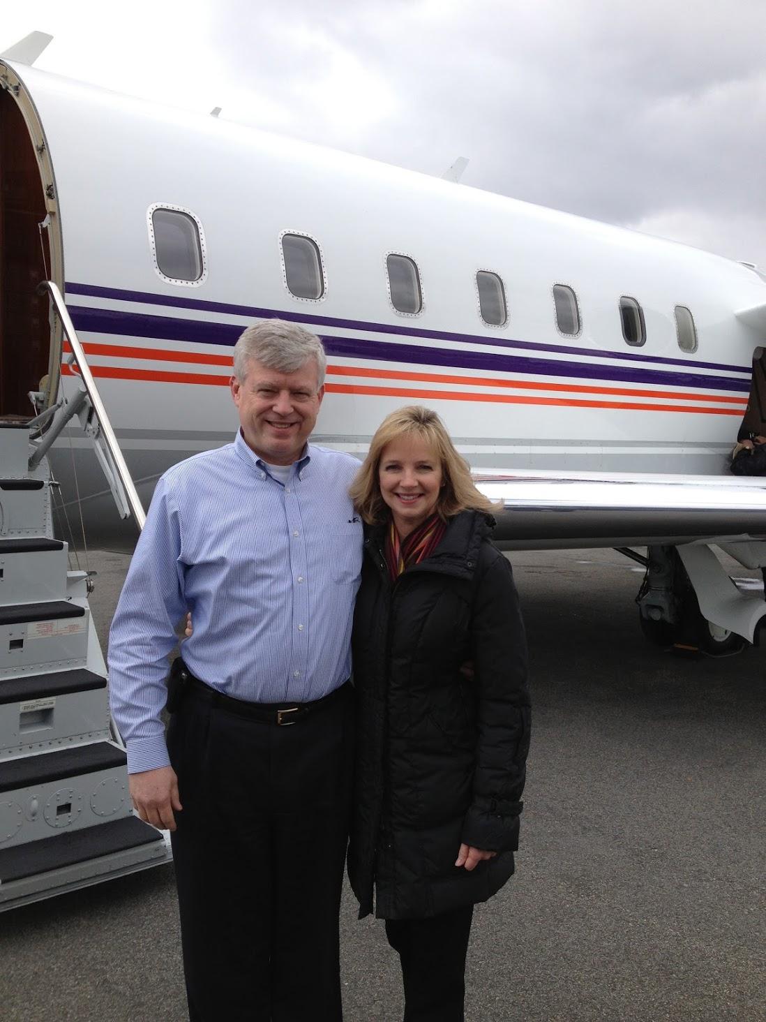 Man in blue shirt and woman in black coat stand smiling in front of a private jet with purple and orange stripes on a cloudy day.