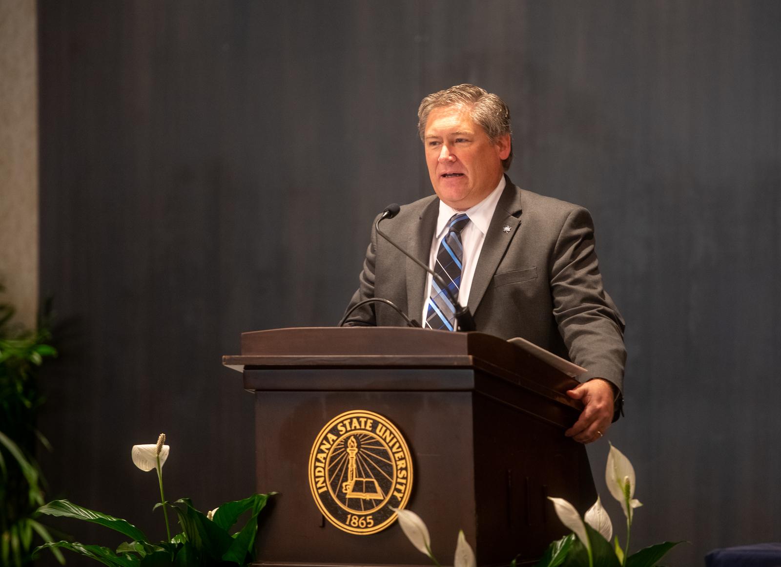 A man in a gray suit and striped tie stands behind a podium with an Indiana State University emblem, speaking into a microphone.
