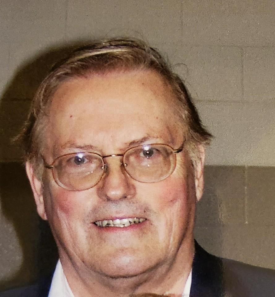 Elderly man with glasses, light hair, and a suit jacket smiling indoors against a plain wall background.