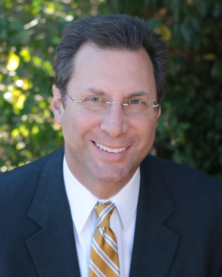 Man wearing glasses, dark suit, white shirt, and yellow striped tie smiling with a blurred green leafy background.