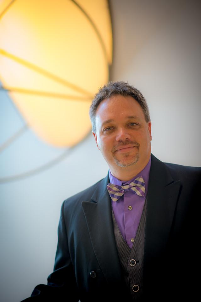 Middle-aged man in a black suit, purple shirt, checkered bow tie, and vest standing indoors with a round glowing light fixture in the background.