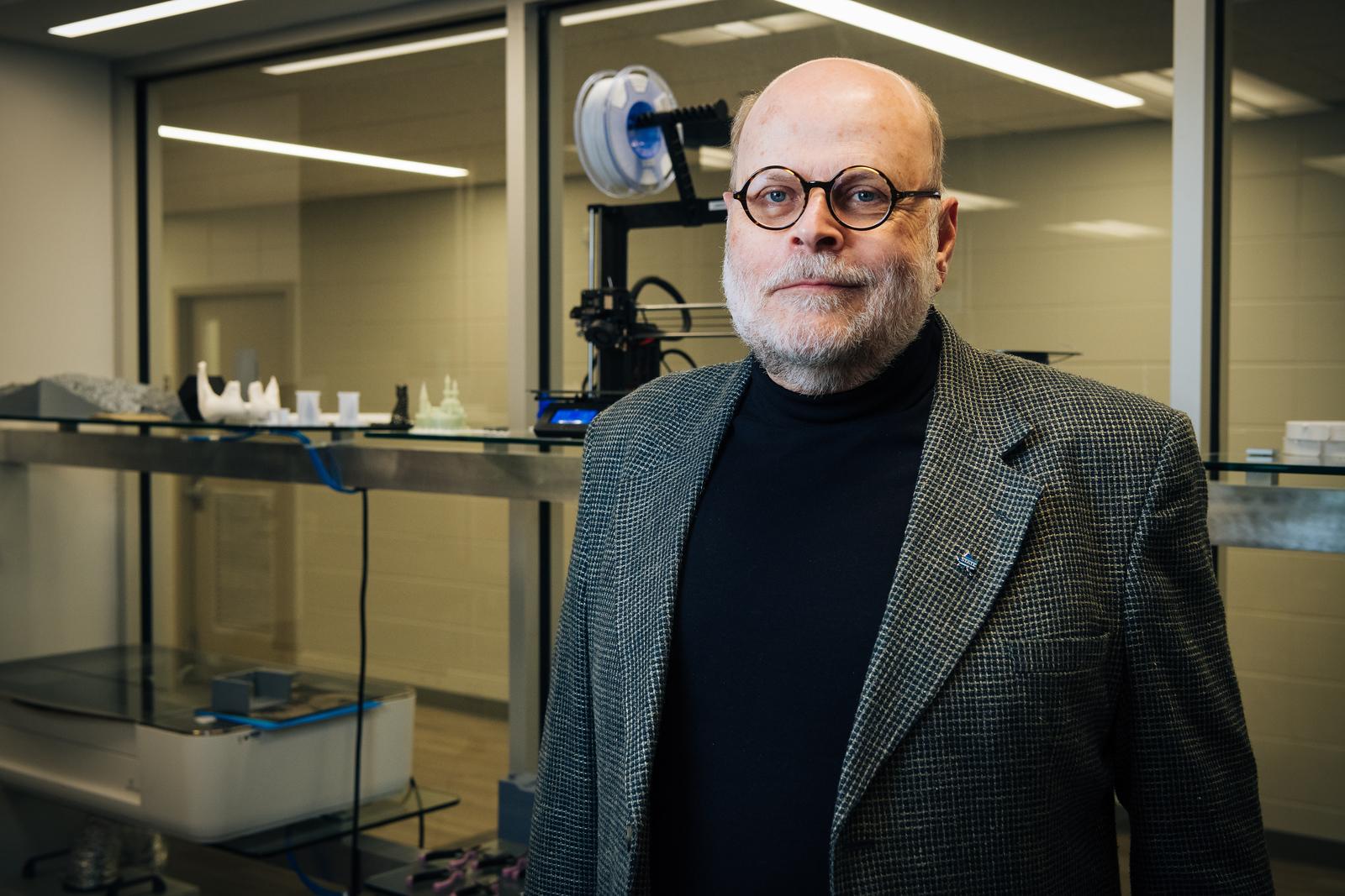 Older man with glasses, beard, and checkered blazer standing in a lab with 3D-printed models and equipment behind him.