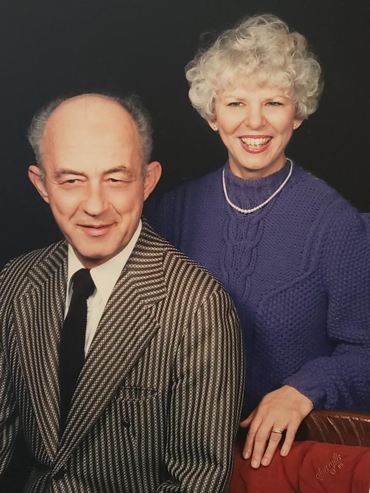 Older man in a striped suit and tie sitting in front of a smiling older woman with curly blonde hair wearing a blue sweater and pearl necklace.