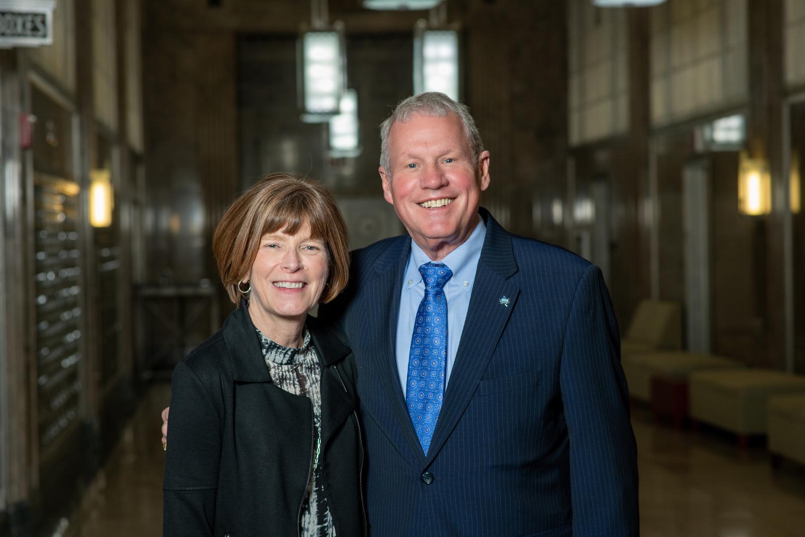 Smiling middle-aged man in a suit and woman in a black jacket standing closely in a dimly lit hallway with lamps and seating in the background.