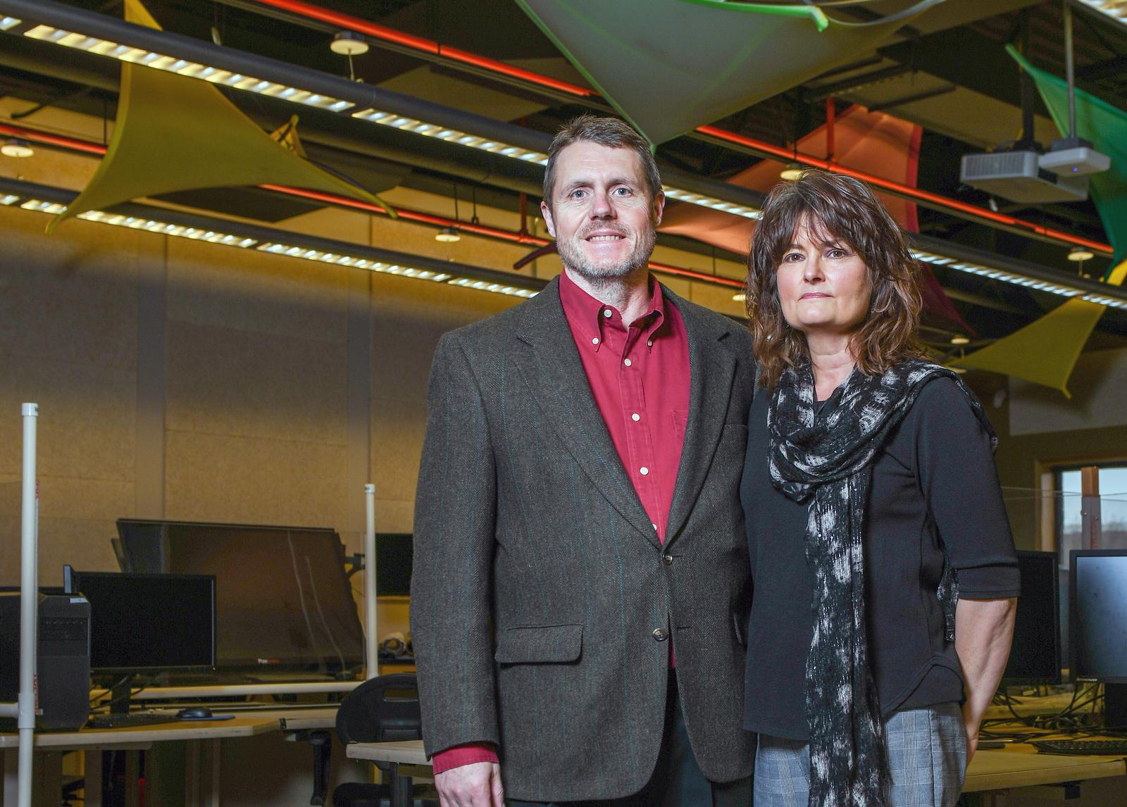 Man in brown blazer and red shirt and woman in black top with scarf stand indoors in a modern computer lab with triangular ceiling decorations.