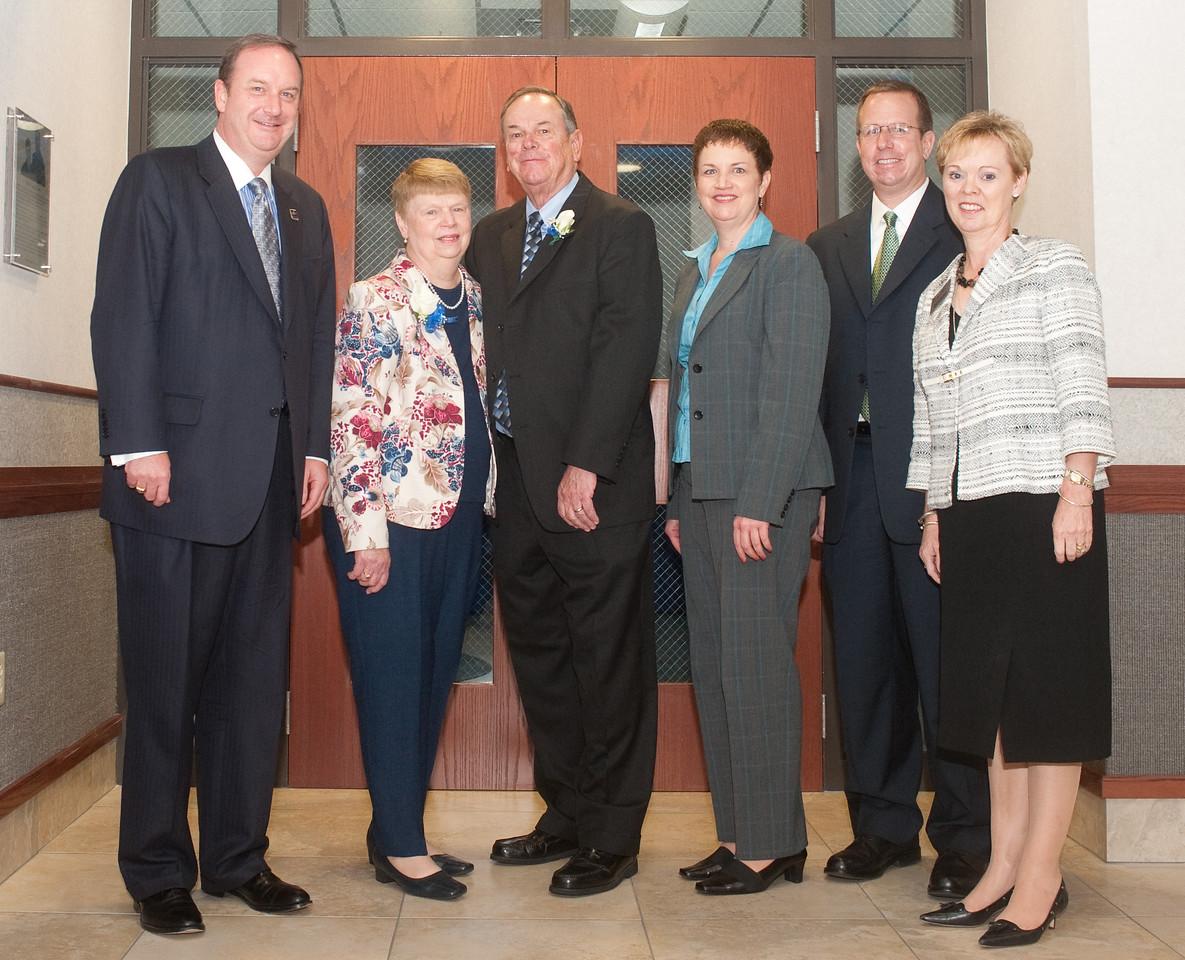 Six people dressed formally stand together smiling in front of a wooden door with glass panels inside a building.