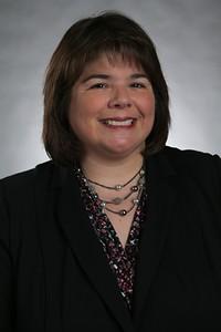 Smiling woman with shoulder-length brown hair wearing a black blazer and layered necklace against a gray background.