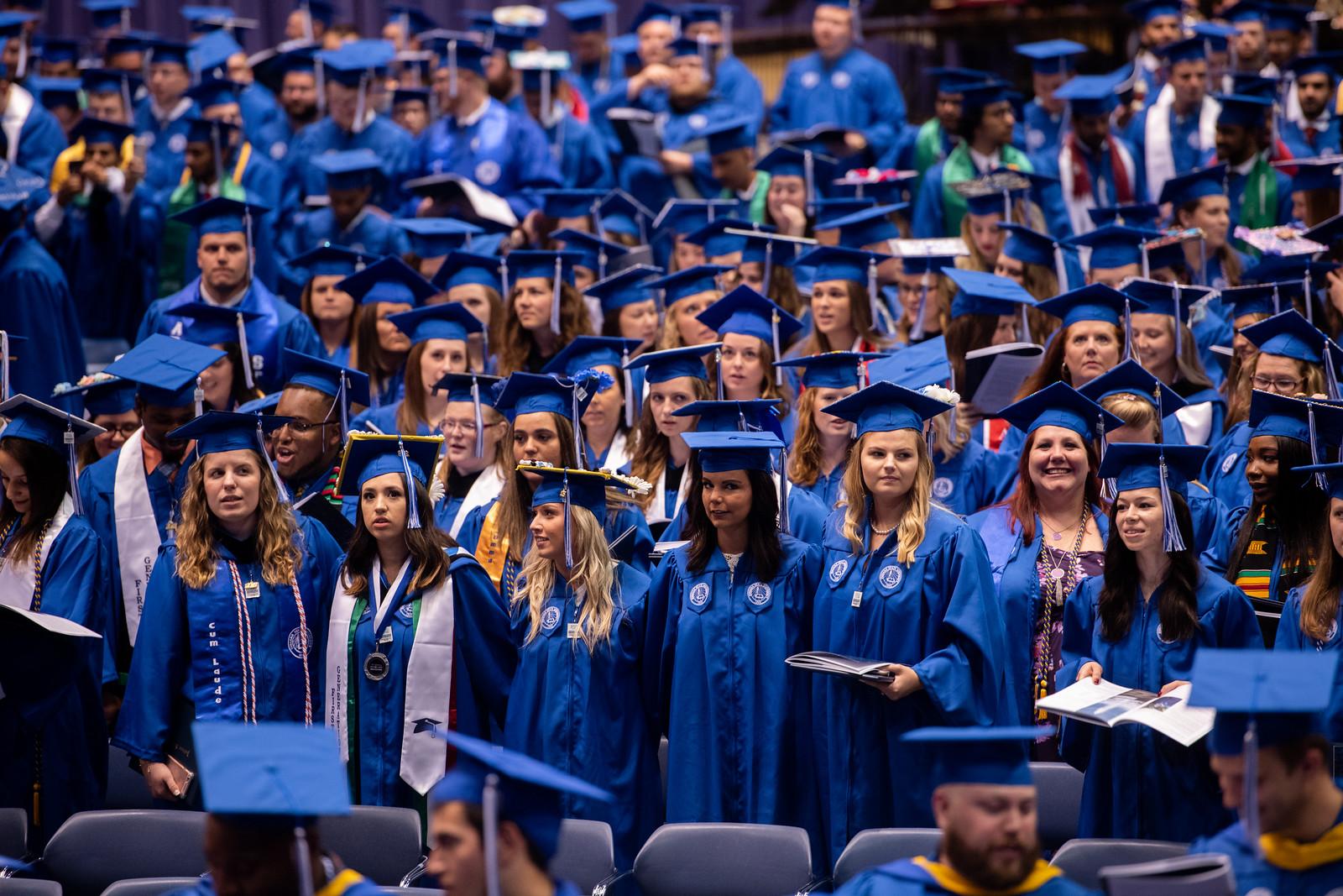 Large group of graduates in blue caps and gowns standing indoors at a commencement ceremony with some holding programs.
