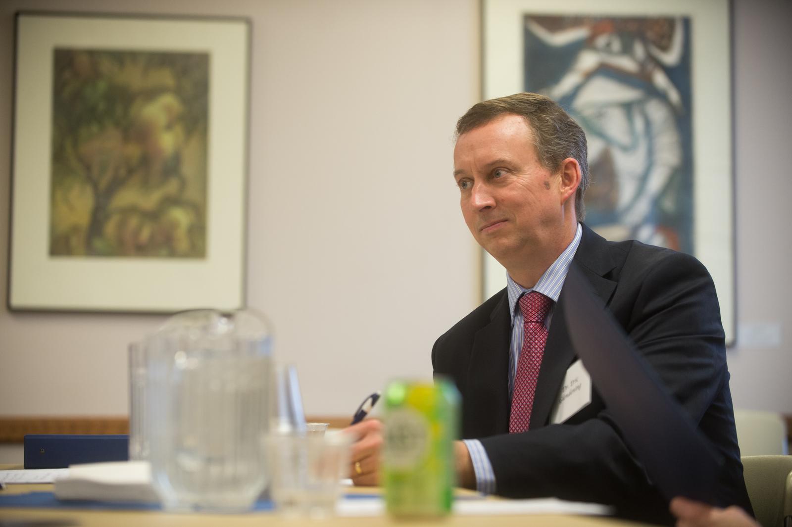Man in suit and red tie sitting at a table with water pitcher, plastic cups, and a soda can, with framed artwork on the wall behind him.