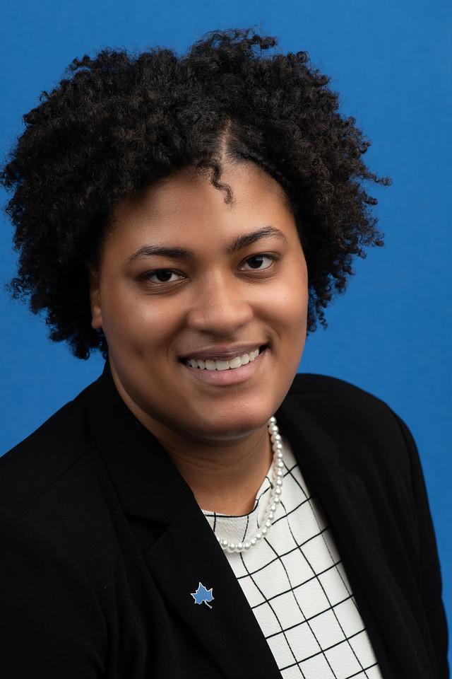 Young woman with curly hair wearing a black blazer, checkered blouse, pearl necklace, and maple leaf pin against a blue backdrop smiling at the camera.