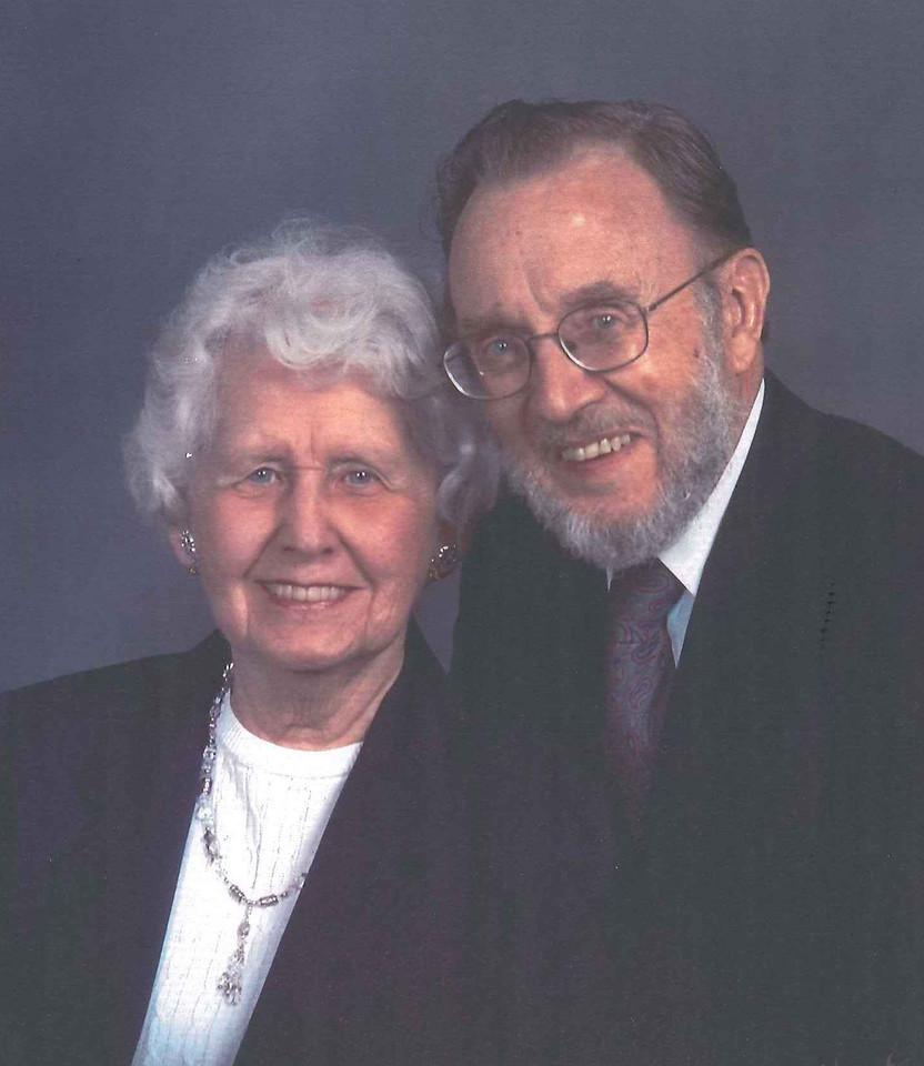 Elderly couple smiling, man with glasses and beard wearing suit and tie, woman with white hair wearing dark jacket and white blouse with necklace.