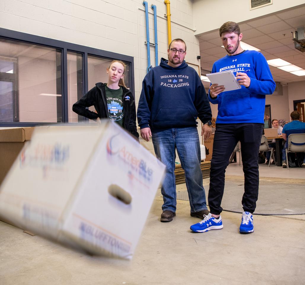 Three students stand in an industrial room examining a tilted cardboard box in front of them, one holding a tablet device.
