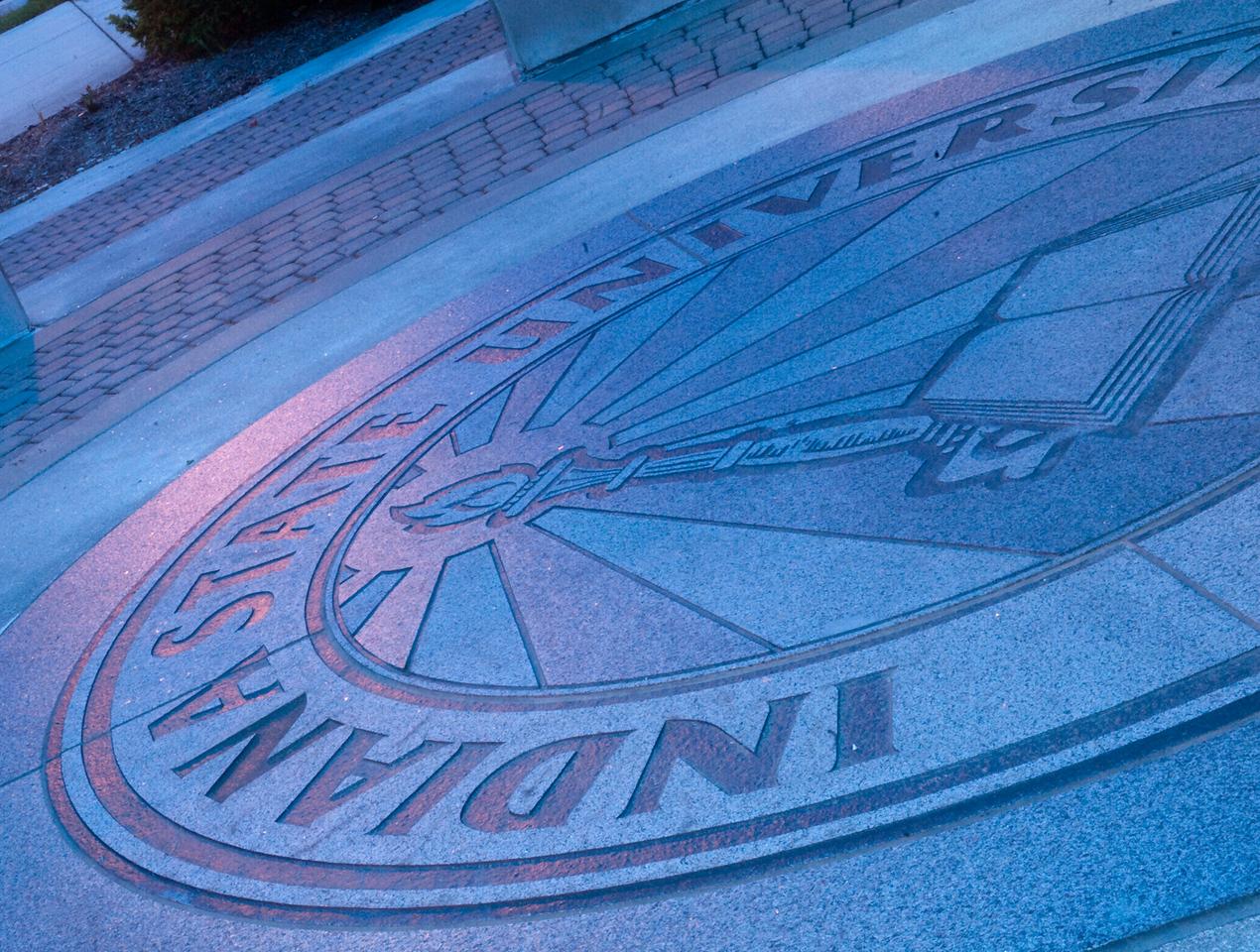 Indiana State University circular emblem on pavement featuring a torch and book design, with landscaping and pavement bricks nearby.