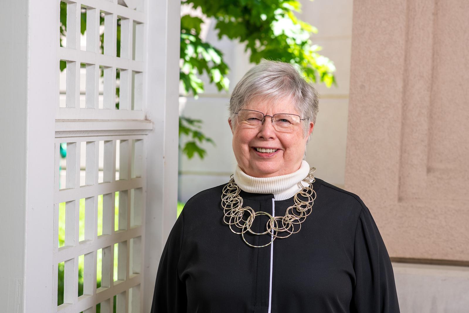 Smiling older woman with short gray hair and glasses wearing a black top and large circular silver necklace standing near a white lattice with greenery behind.