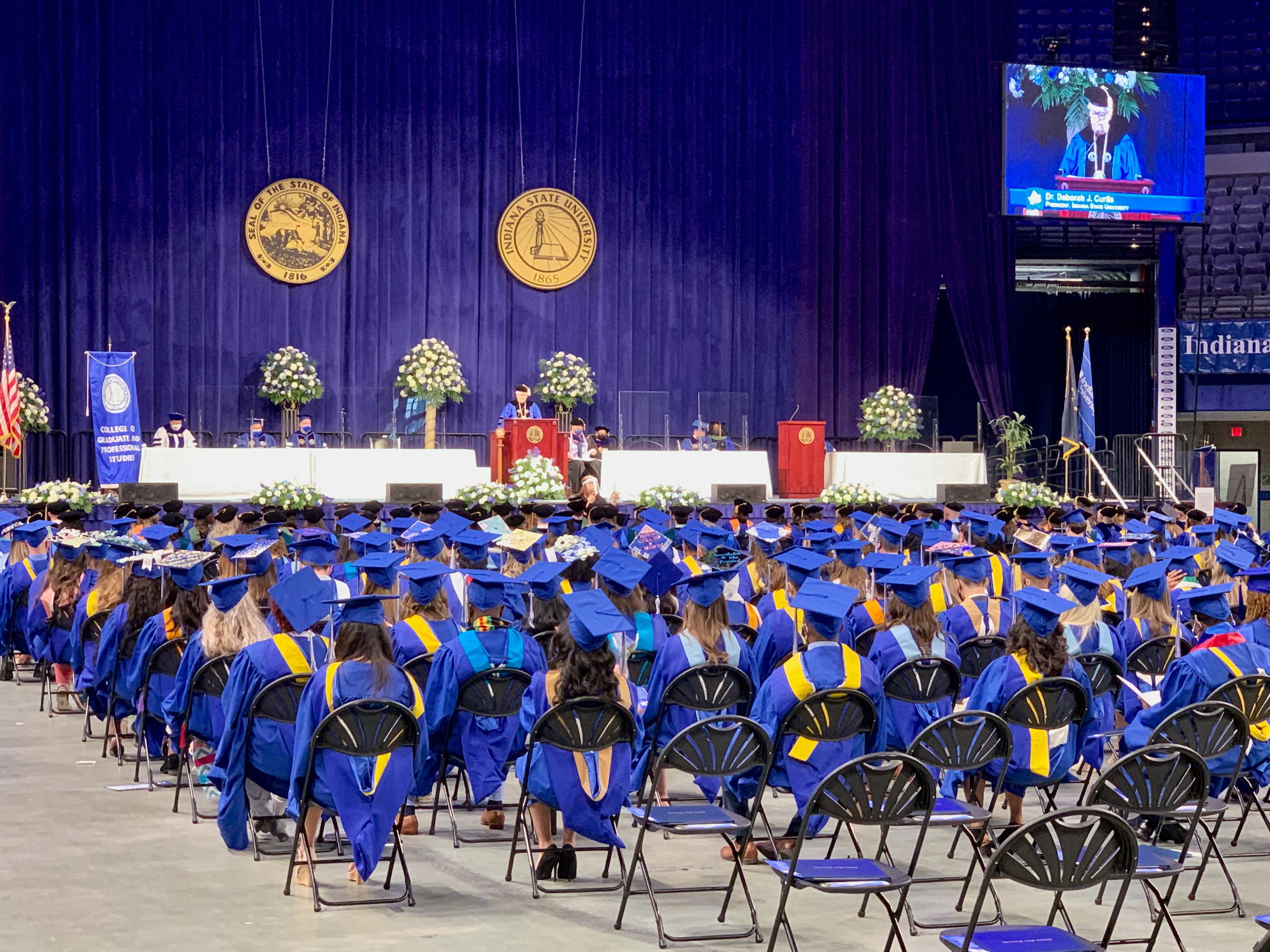 Graduates in blue caps and gowns seated facing a stage with speakers and floral arrangements in a large hall during graduation ceremony.