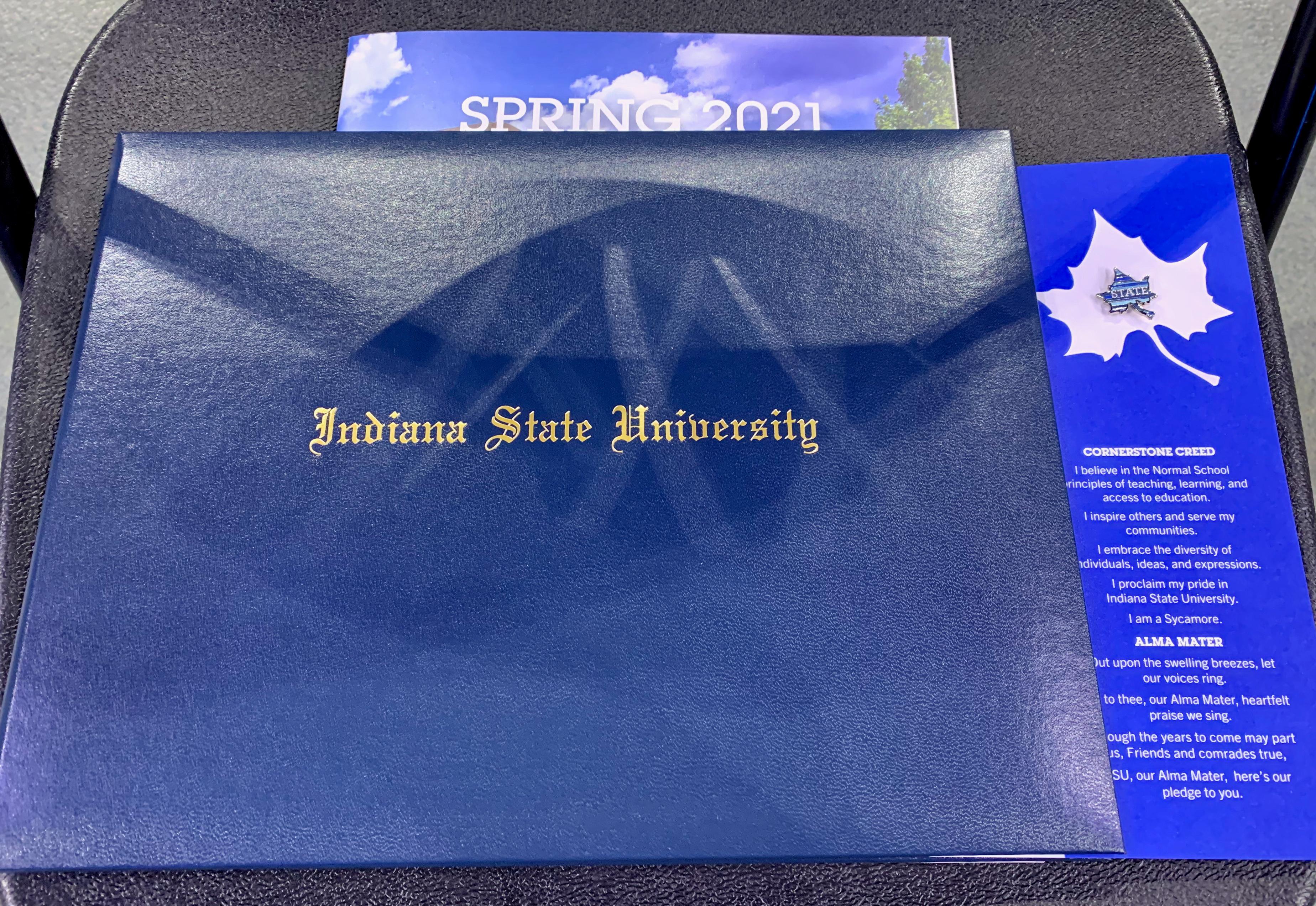 Blue diploma cover with gold Indiana State University text, partially covered Spring 2021 program, and a blue card with university pin and Creed text.