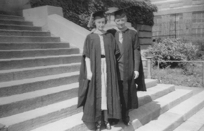 Two graduates in caps and gowns stand on stone steps outside a large building with trees and shrubs nearby.