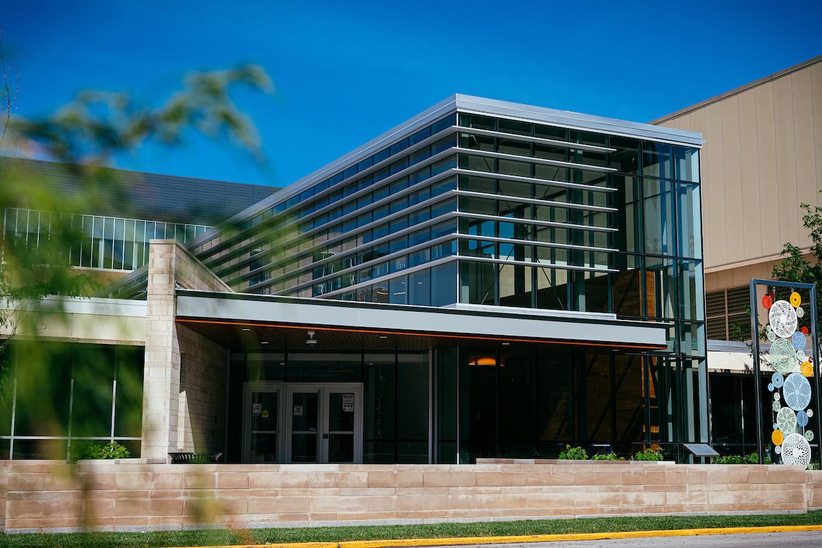 Modern glass and stone university building under blue sky with outdoor decorative art installation on the right side of the entrance.