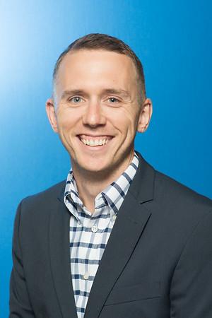 Smiling man with short hair wearing a checkered shirt and dark blazer against a blue background