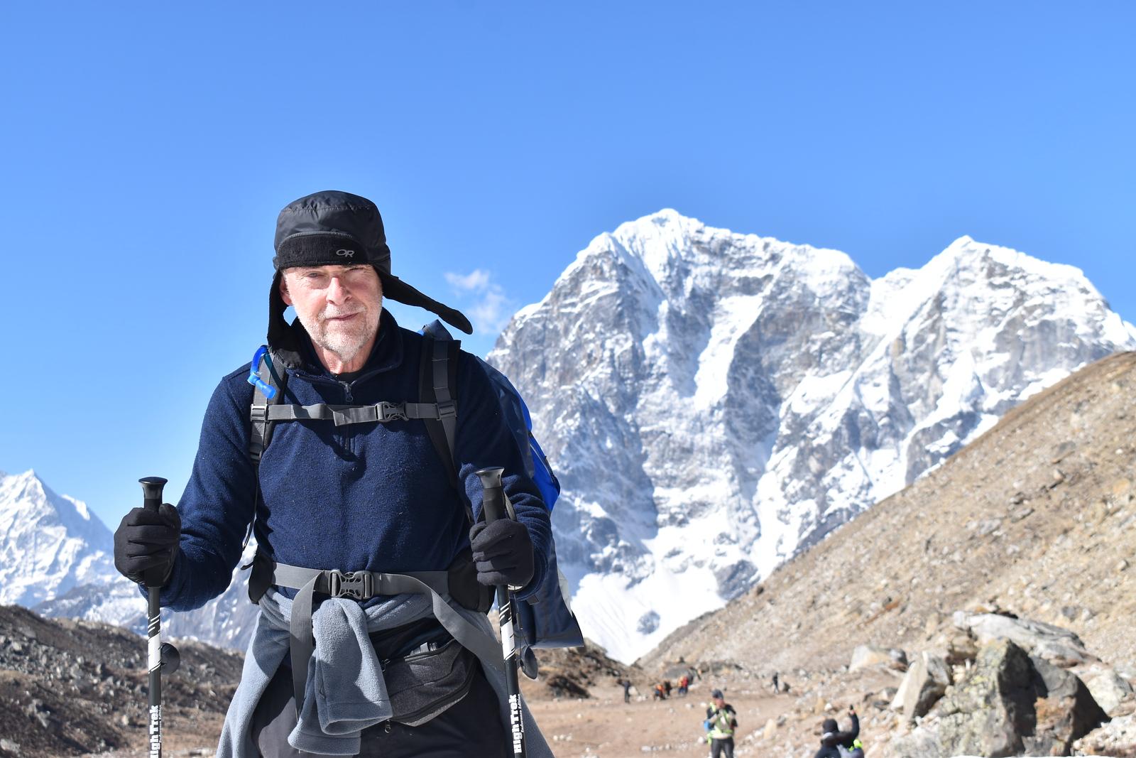 Older man wearing winter hiking gear and holding trekking poles stands on rocky mountain trail with snowy peaks in the background.
